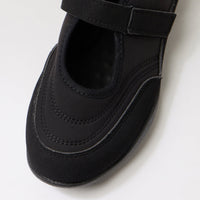 A close-up of a black Mary Jane style shoe with a hook-and-loop strap, photographed on a clean white background.